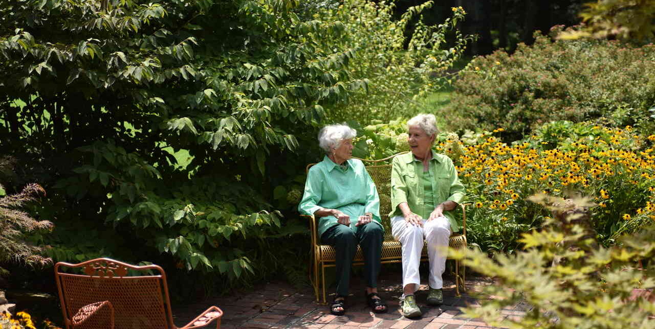 2 women sitting in the middle of the garden surrounded by flowers and greenery